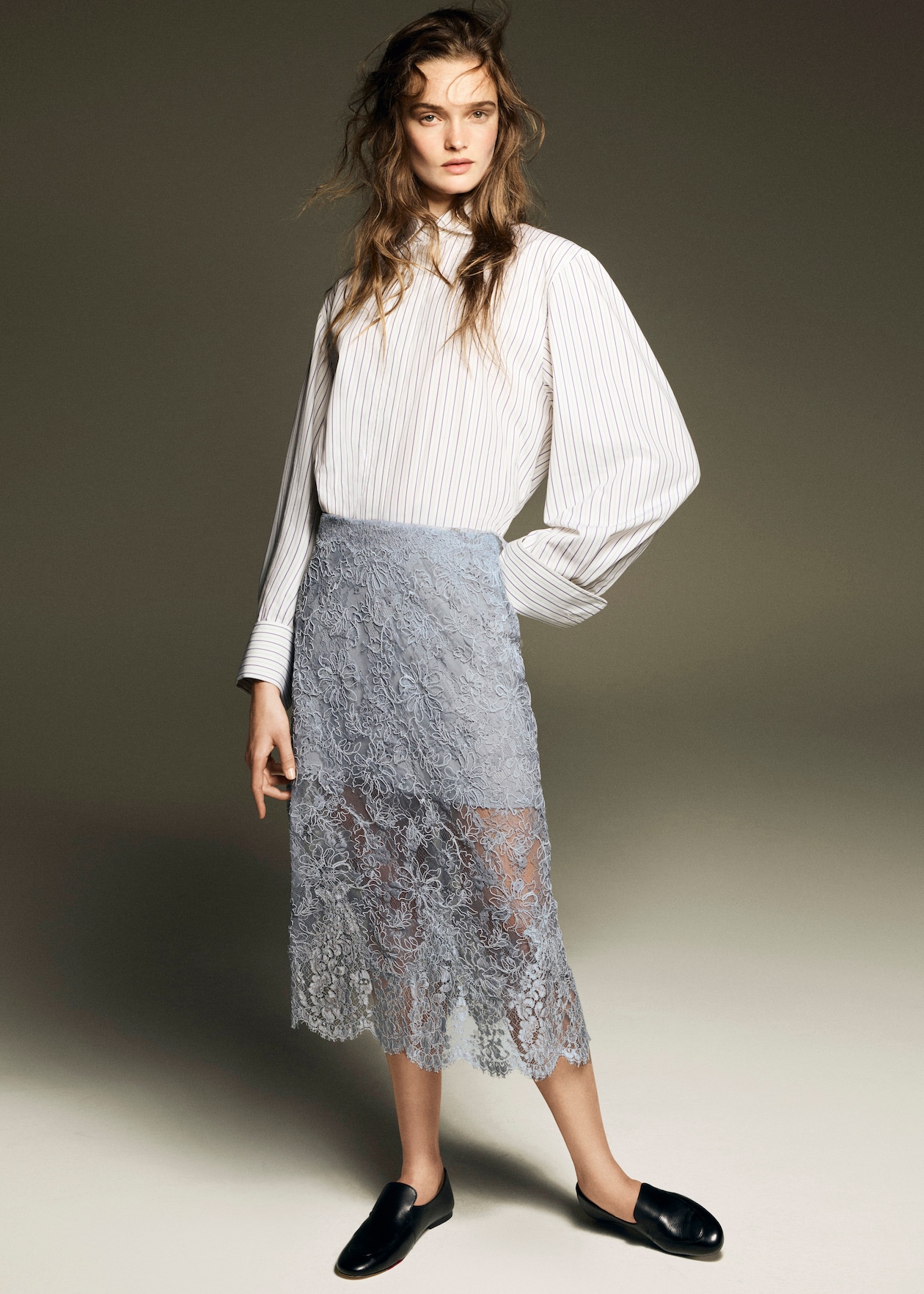 A woman in a striped blouse and sheer lace skirt, with long hair, poses against a neutral background.