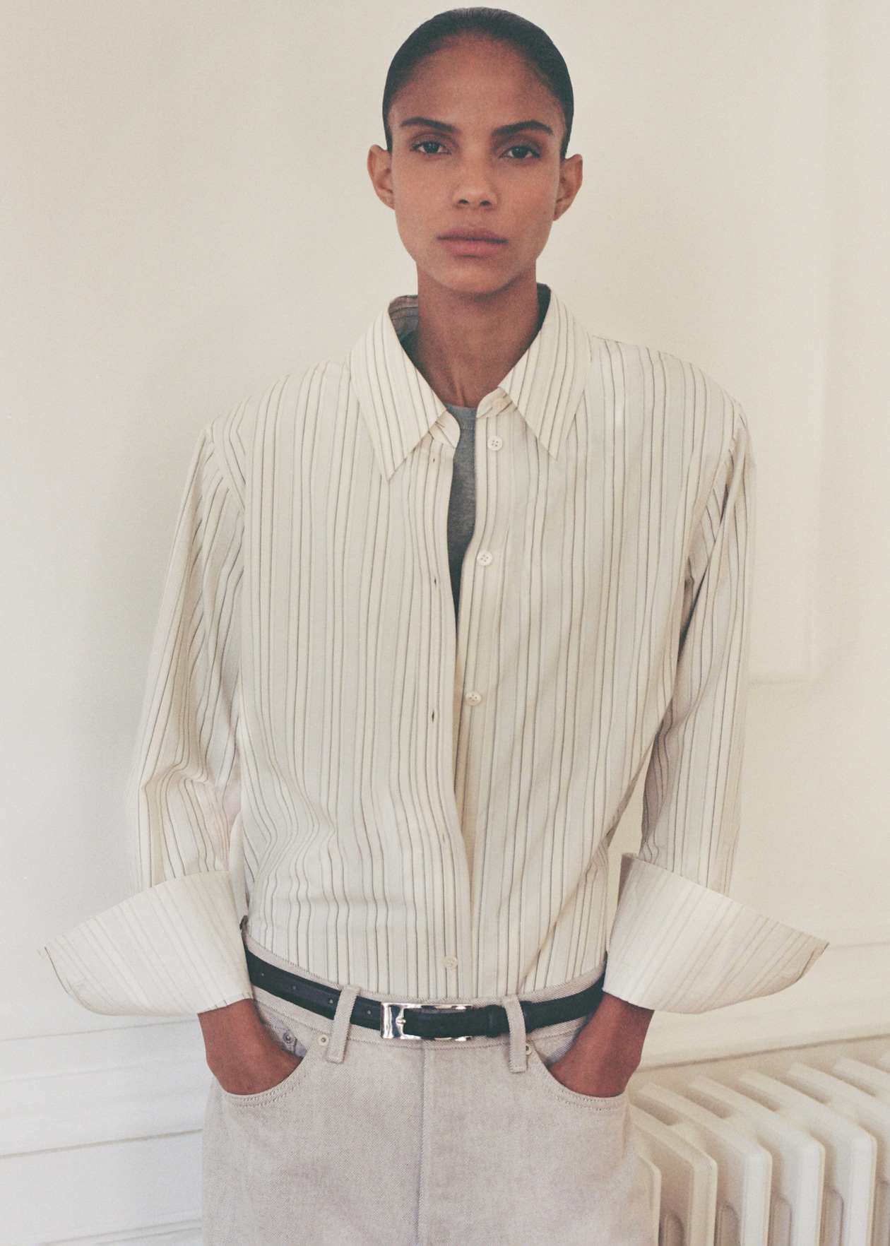 Person wearing a loose, striped button-up shirt with rolled sleeves, hands in pockets, standing against a light-colored wall.