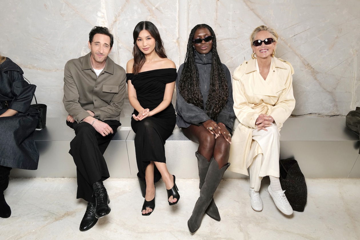 Four people seated on a bench, dressed in stylish, modern attire, posing for a photo against a light-colored background.