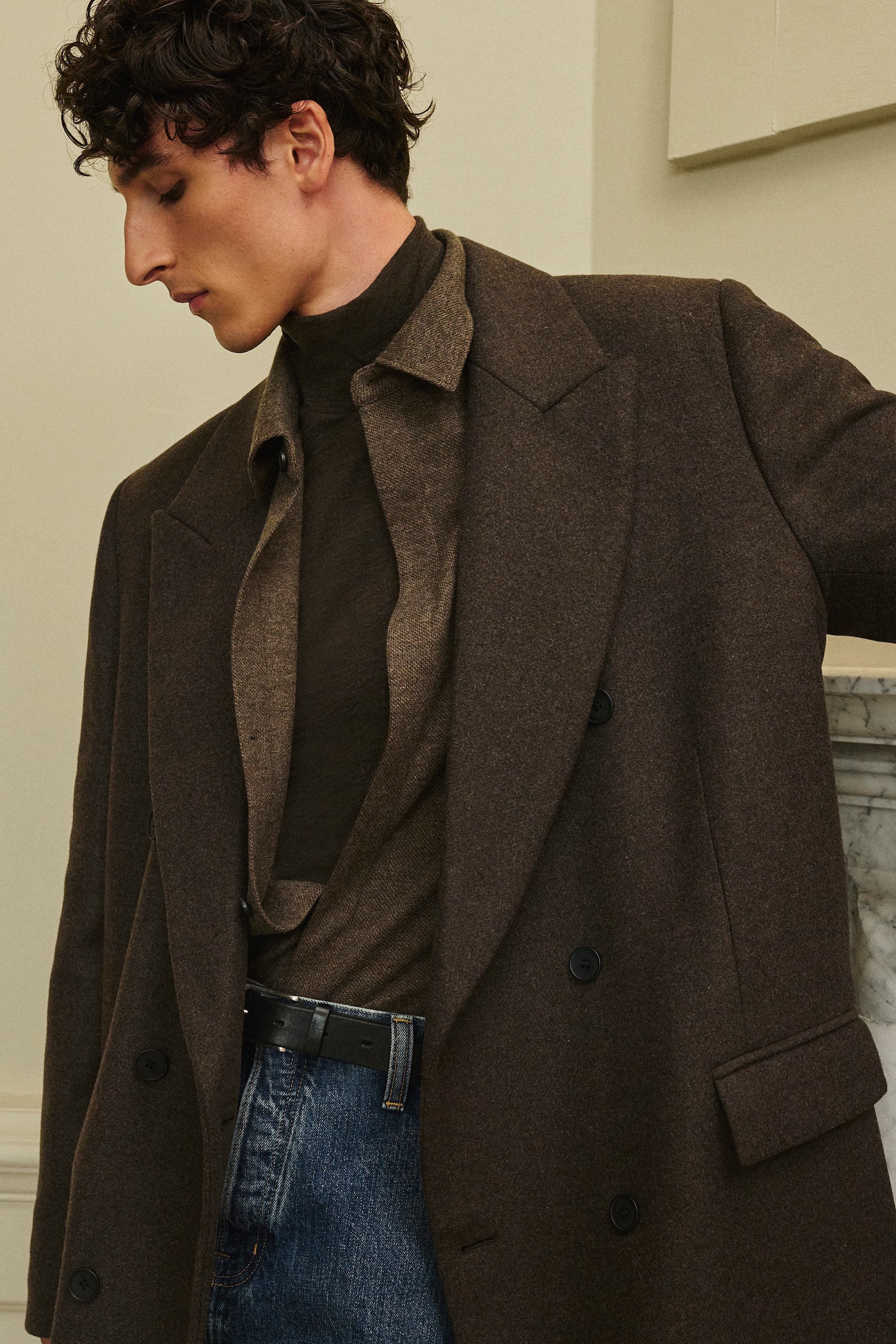 A person wearing a brown coat over a brown turtleneck and shirt, paired with blue jeans, stands against a neutral background.