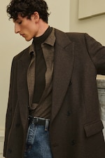 Placeholder for: A person wearing a brown coat over a brown turtleneck and shirt, paired with blue jeans, stands against a neutral background.