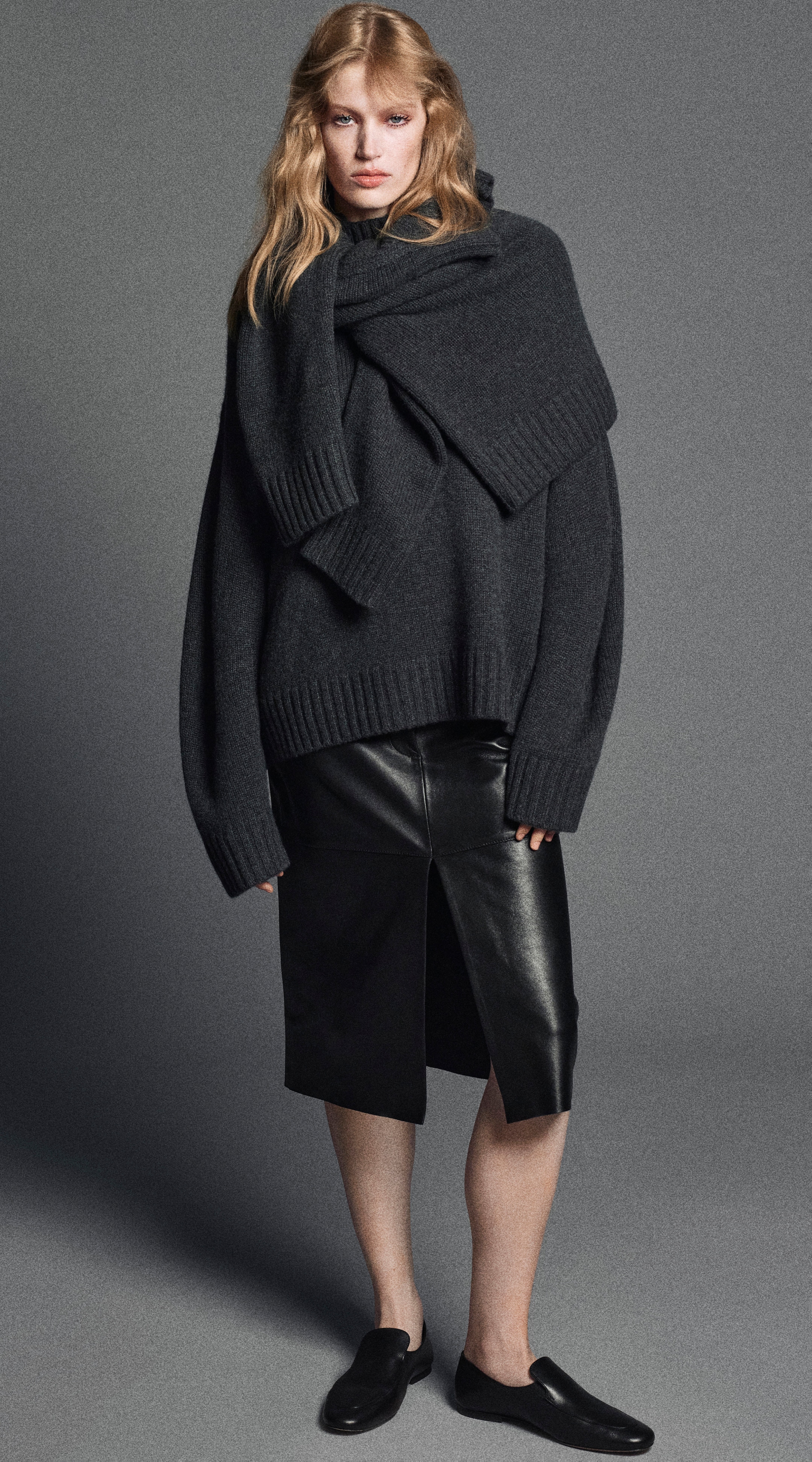 Woman wearing an oversized dark sweater, black leather skirt, and black shoes, posing against a gray background.