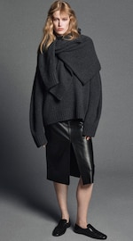 Placeholder for: Woman wearing an oversized dark sweater, black leather skirt, and black shoes, posing against a gray background.