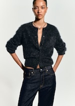 Placeholder for: A woman wearing a dark, fluffy cardigan and blue jeans stands confidently, her hair tied back.