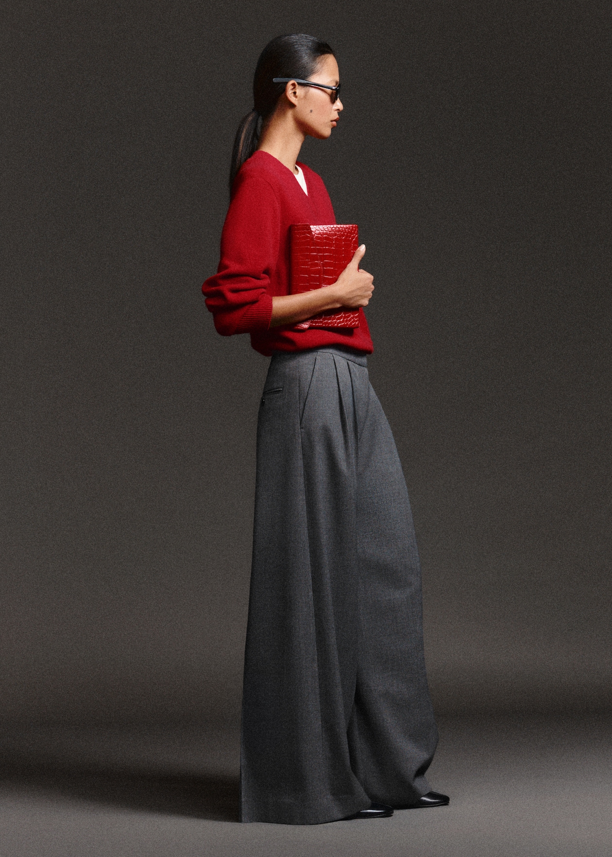 A person in a red sweater and gray wide-leg pants holds a red clutch and wears glasses against a dark background.