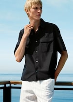 Placeholder for: A person with blond hair wearing a black button-up shirt and white pants stands near a railing with a clear blue sky and sea in the background.