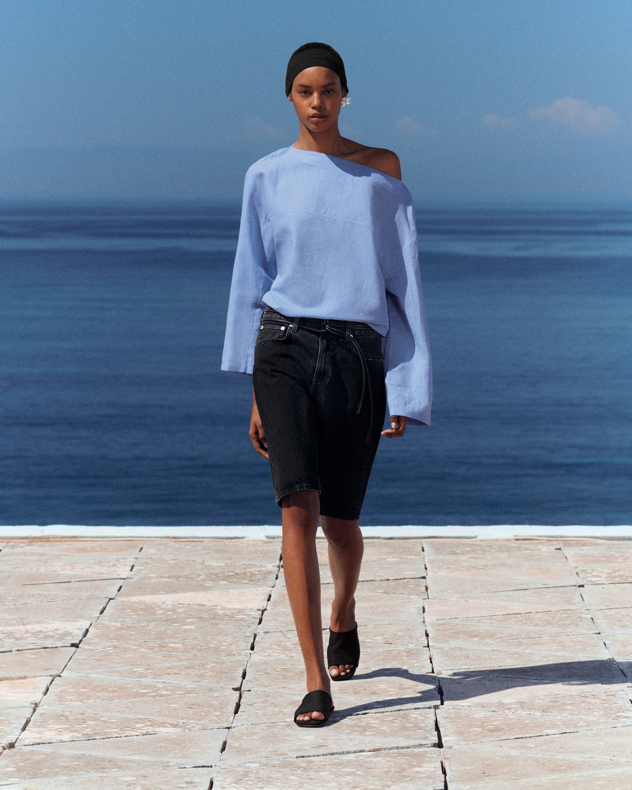 Person walking outdoors by the sea, wearing a light blue off-shoulder top, dark shorts, black sandals, and a headband, with a serene ocean backdrop.