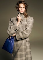 Placeholder for: A person wearing a checkered coat holds a blue handbag, with a thoughtful expression, in front of a neutral background.