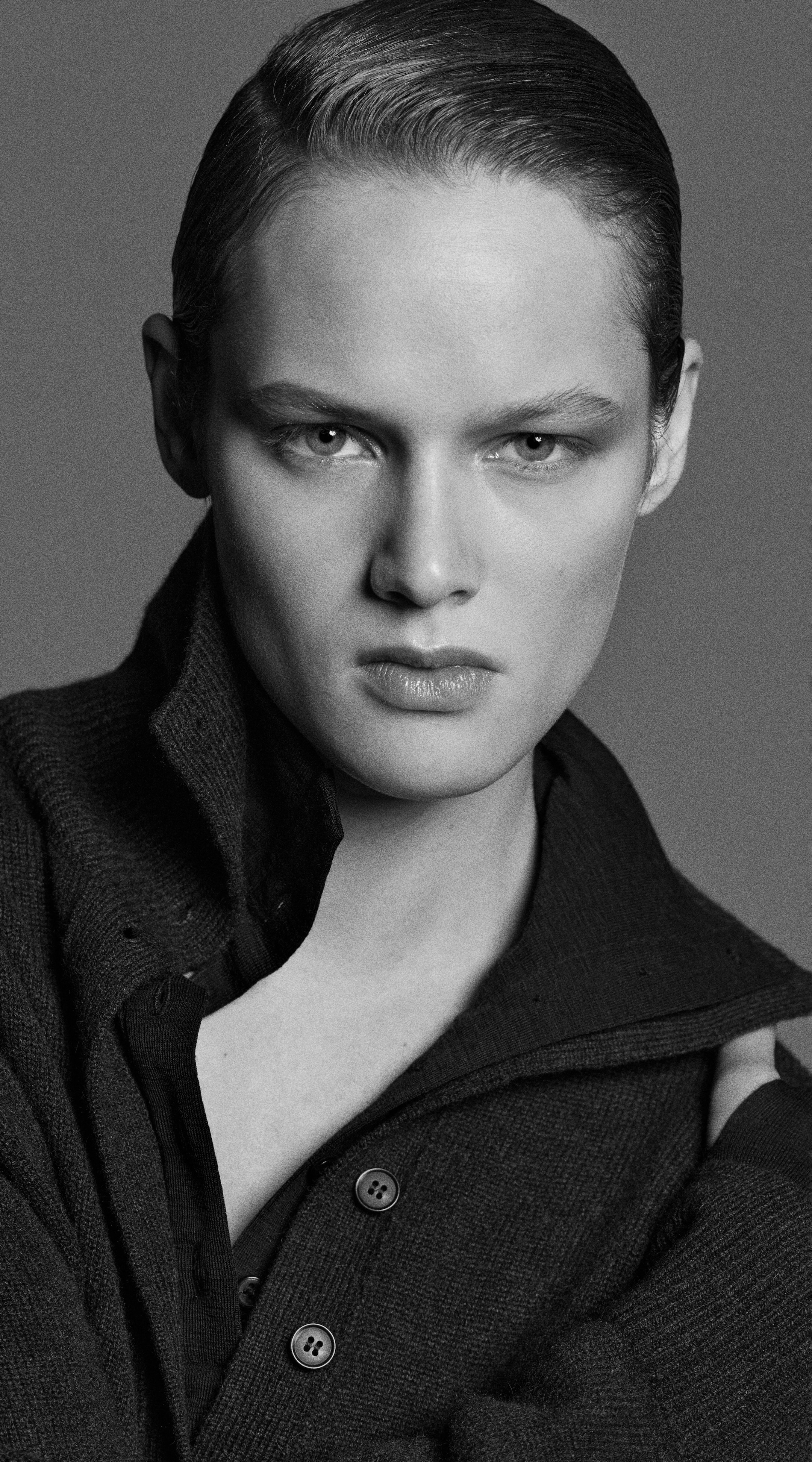 Black and white portrait of a person with slicked-back hair, wearing a dark buttoned sweater, looking intensely at the camera.