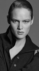 Placeholder for: Black and white portrait of a person with slicked-back hair, wearing a dark buttoned sweater, looking intensely at the camera.