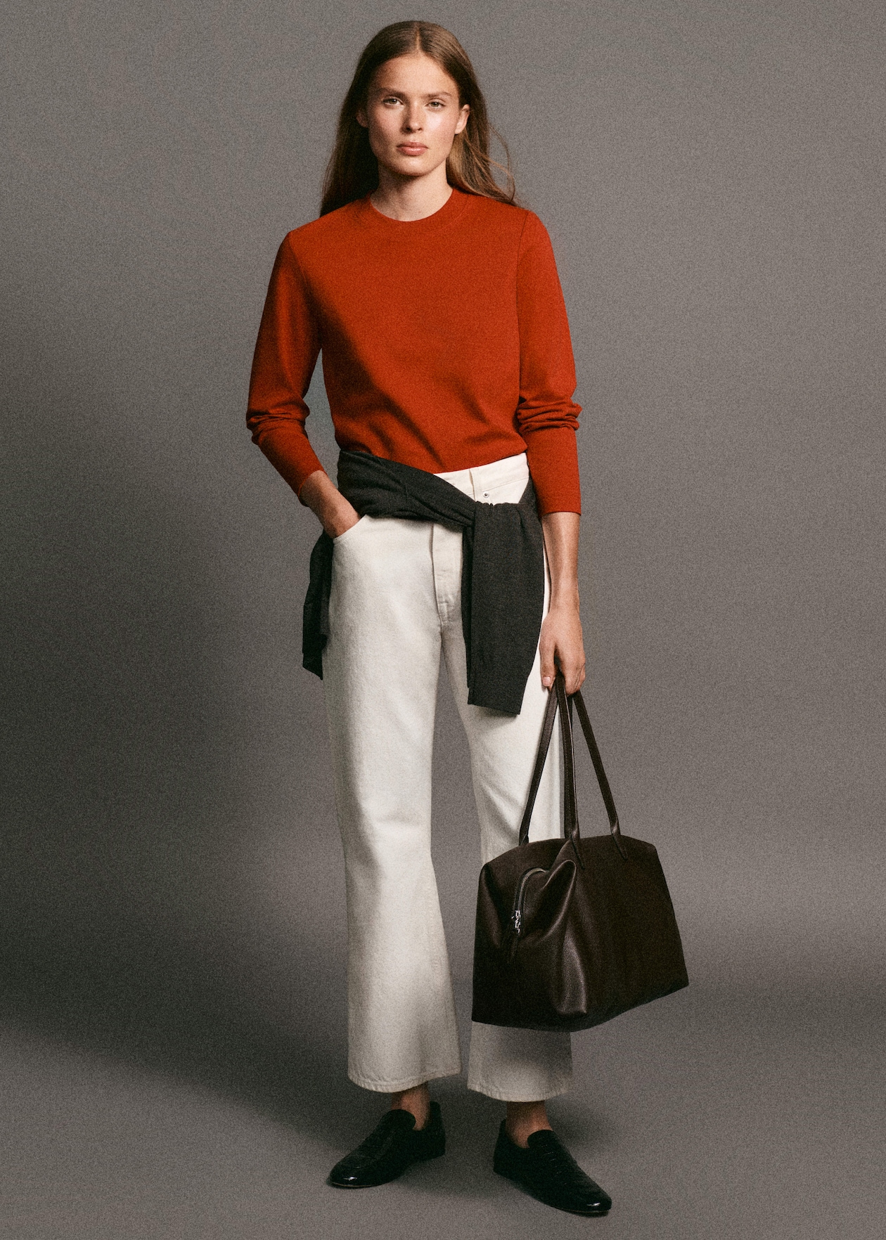 A person wearing a red sweater, white pants, and black loafers holds a dark brown tote bag, with a black sweater tied around the waist.