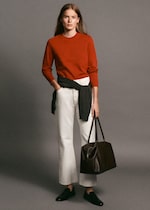 Placeholder for: A person wearing a red sweater, white pants, and black loafers holds a dark brown tote bag, with a black sweater tied around the waist.
