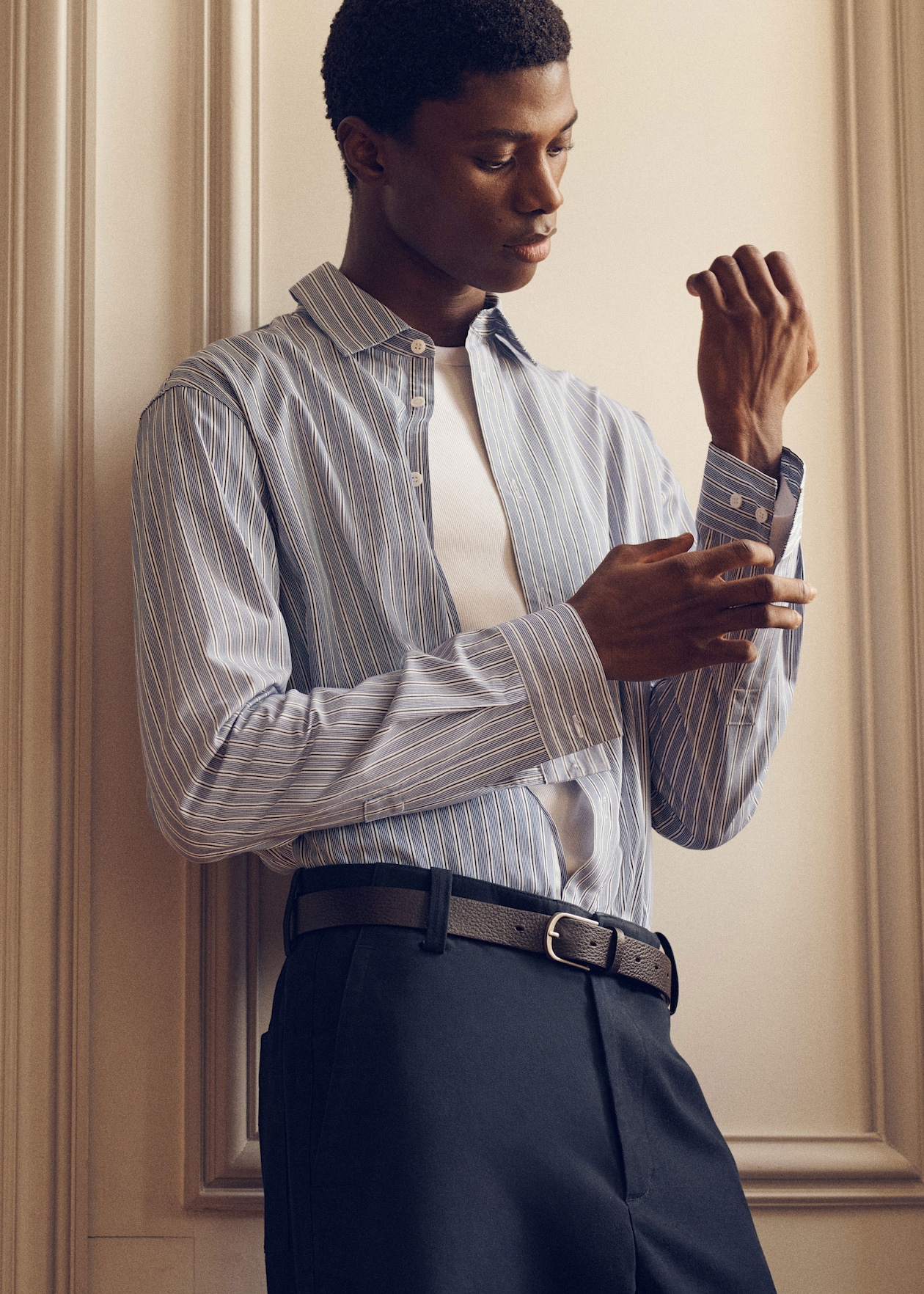 A man adjusts the cuff of his striped blue shirt, wearing a white undershirt and dark trousers, standing against a cream-colored wall.