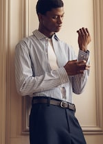 Placeholder for: A man adjusts the cuff of his striped blue shirt, wearing a white undershirt and dark trousers, standing against a cream-colored wall.