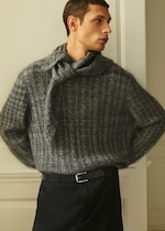 Placeholder for: Person wearing a grey textured sweater with a scarf tied around the neck, looking to the side, standing in a well-lit room.