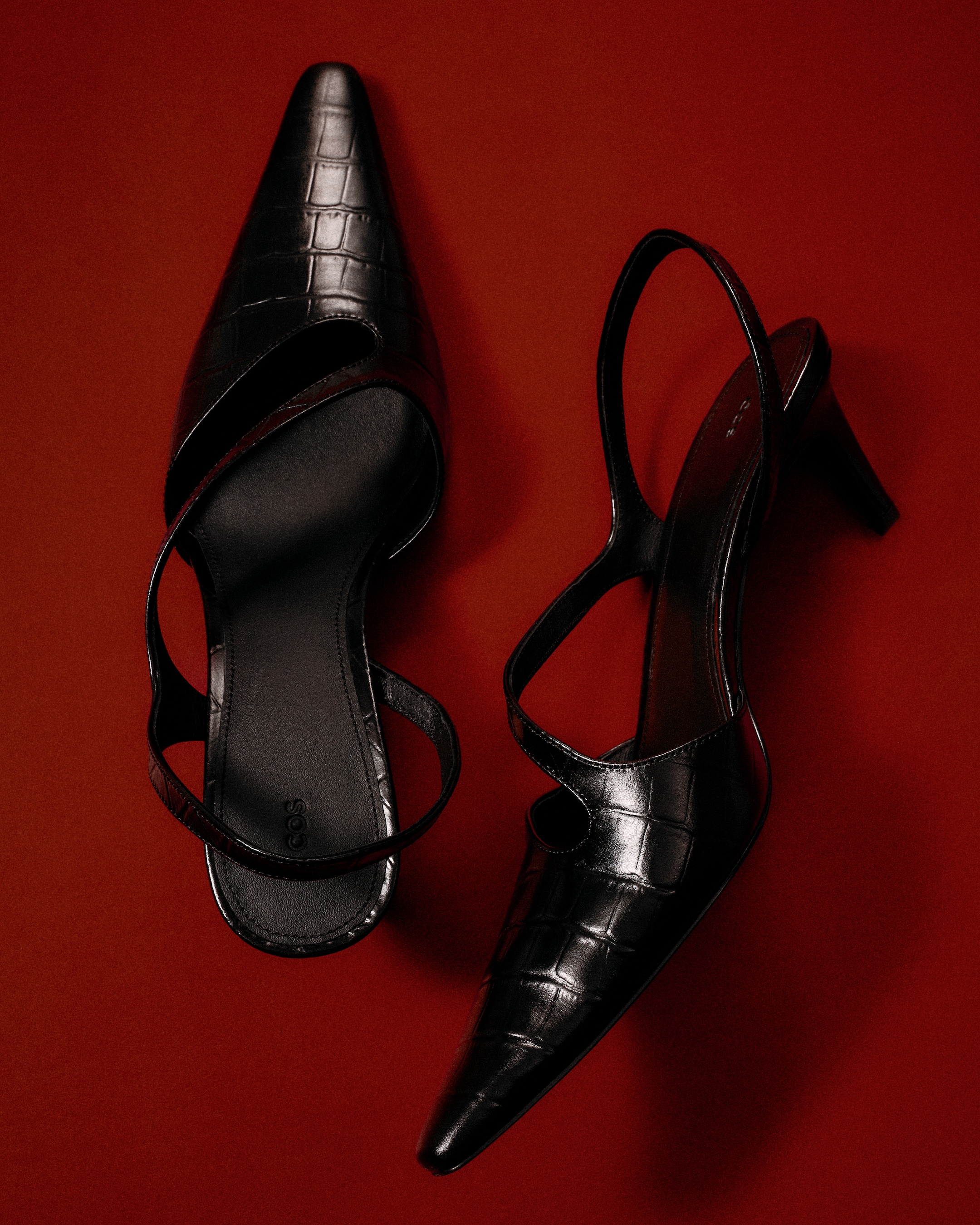 Pair of black, pointed-toe high heels with a crocodile texture on a red background.
