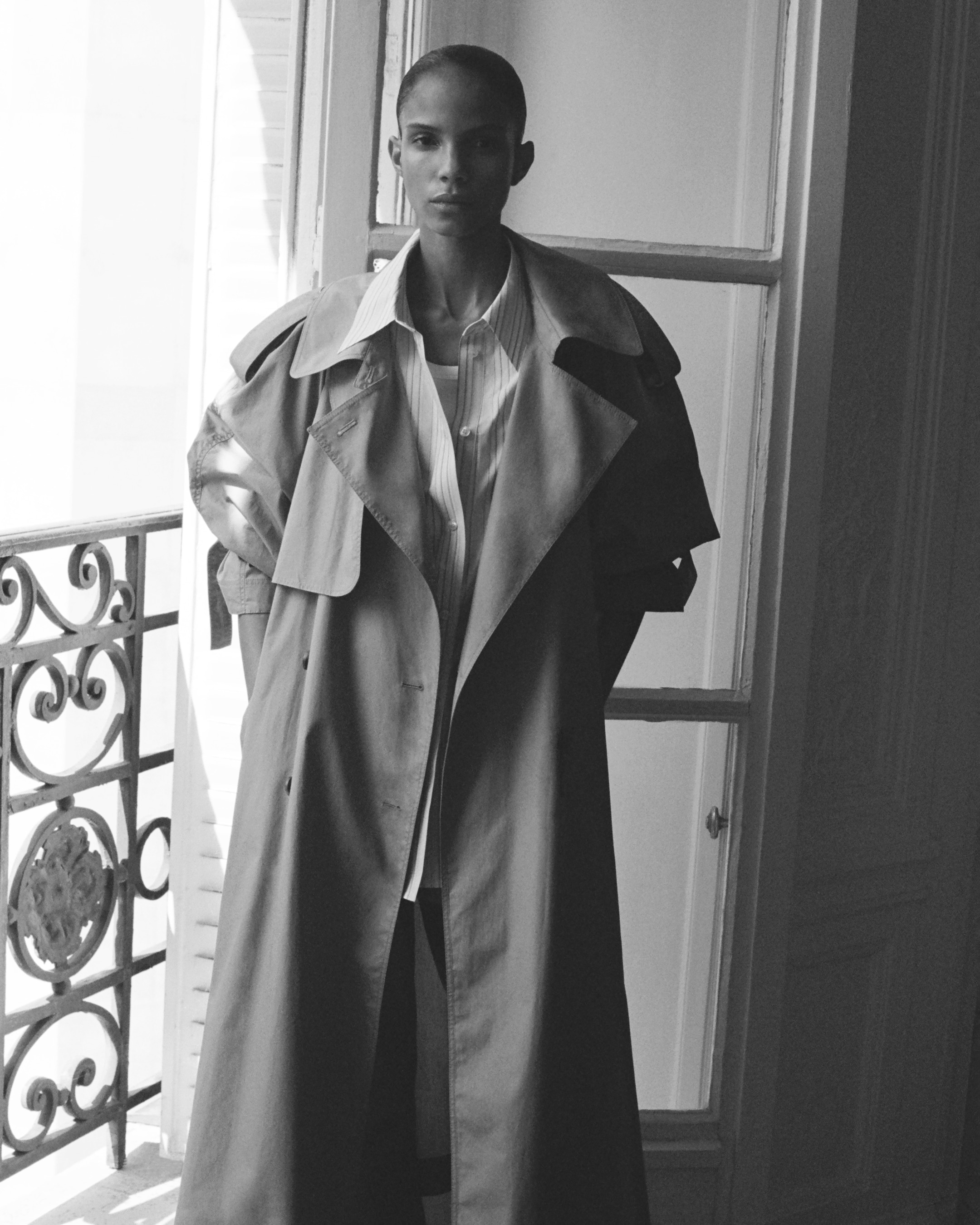 A person stands in a doorway, wearing an oversized trench coat and a light shirt, with a decorative railing visible on the left.