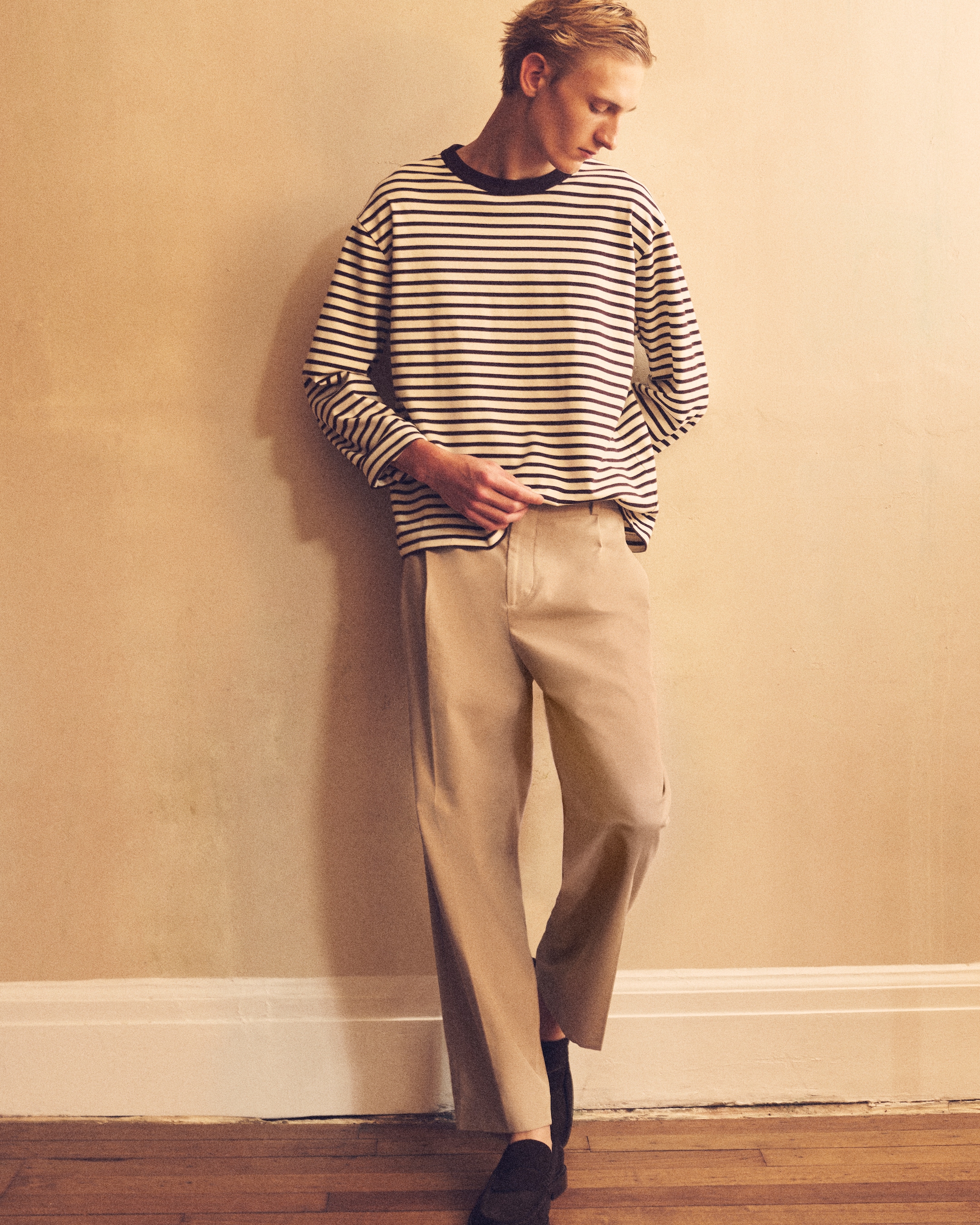 Man standing against a beige wall, with brown trousers on, black shoes on and a striped long sleeve t-shirt on