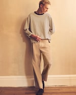 Placeholder for: Man standing against a beige wall, with brown trousers on, black shoes on and a striped long sleeve t-shirt on