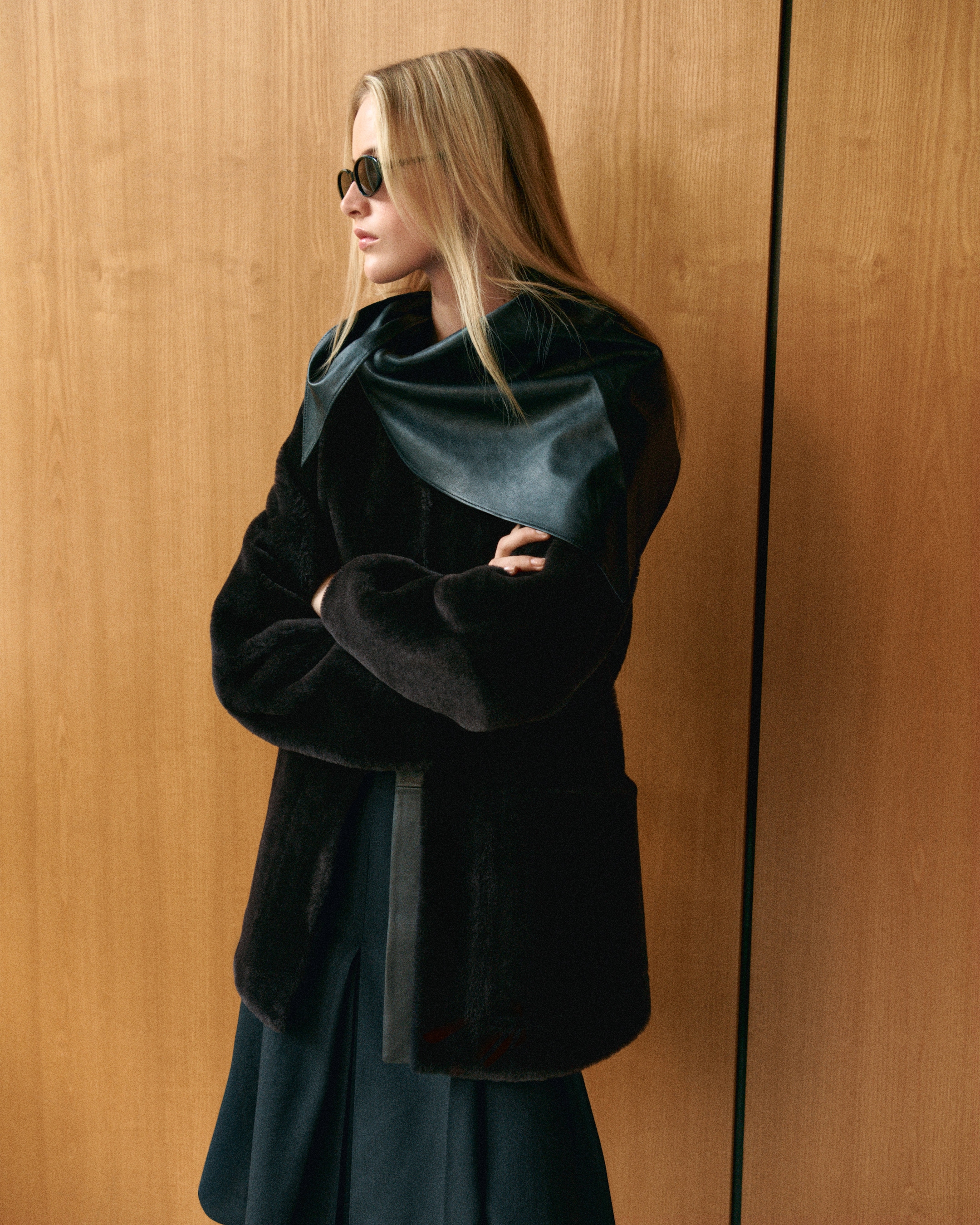 A woman in sunglasses and a dark winter coat stands against a wooden wall, looking to the side with arms crossed.