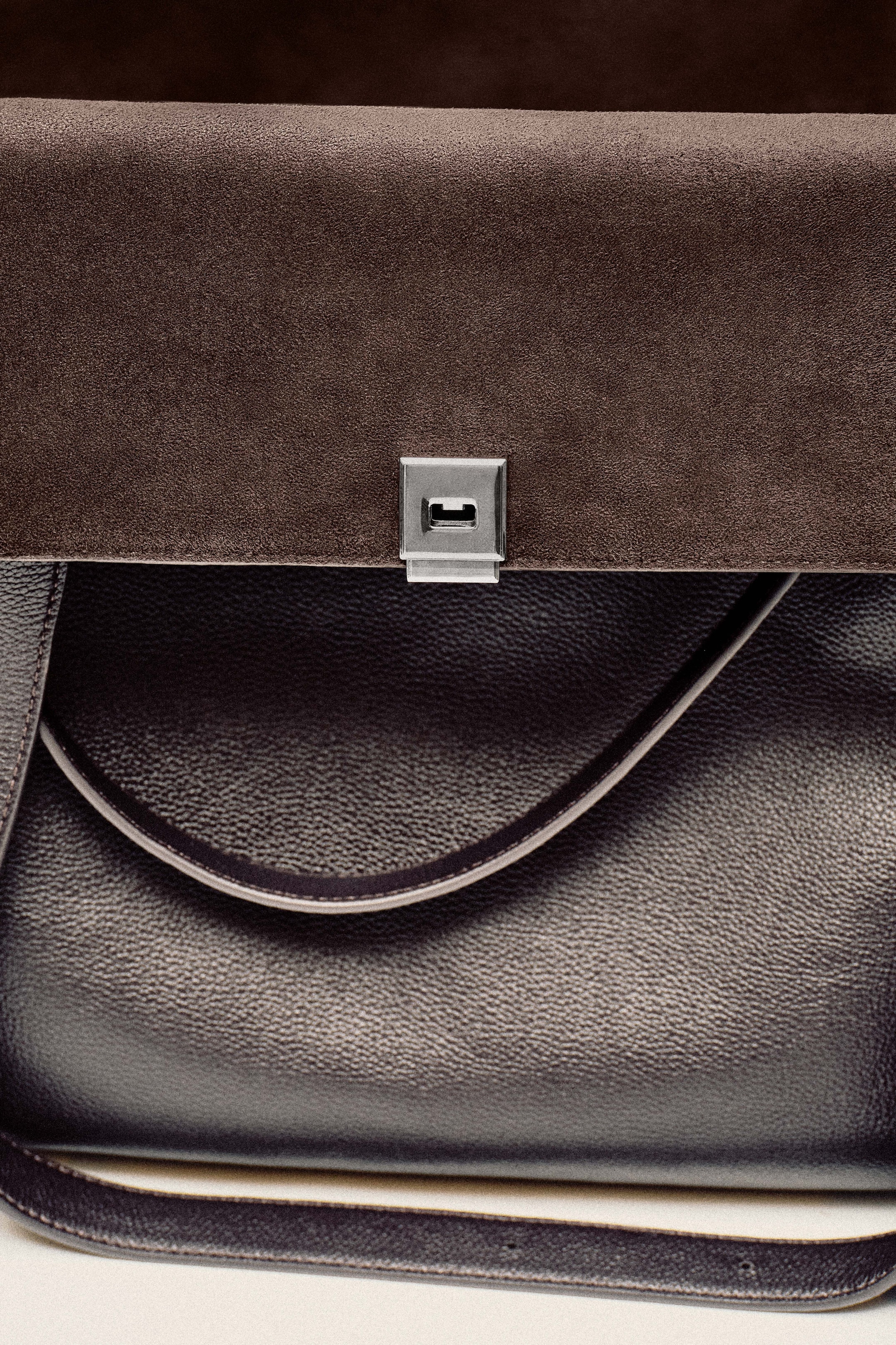 Close-up of a luxury handbag with a dark leather body and a brown suede flap, featuring a silver lock.