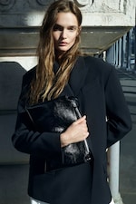 Placeholder for: A woman with long hair wearing a black blazer holds a black leather clutch, standing against a textured wall, outdoors in sunlight.