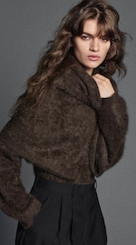Placeholder for: A person with long brown hair wearing a dark, textured sweater and black pants, posing against a gray background.