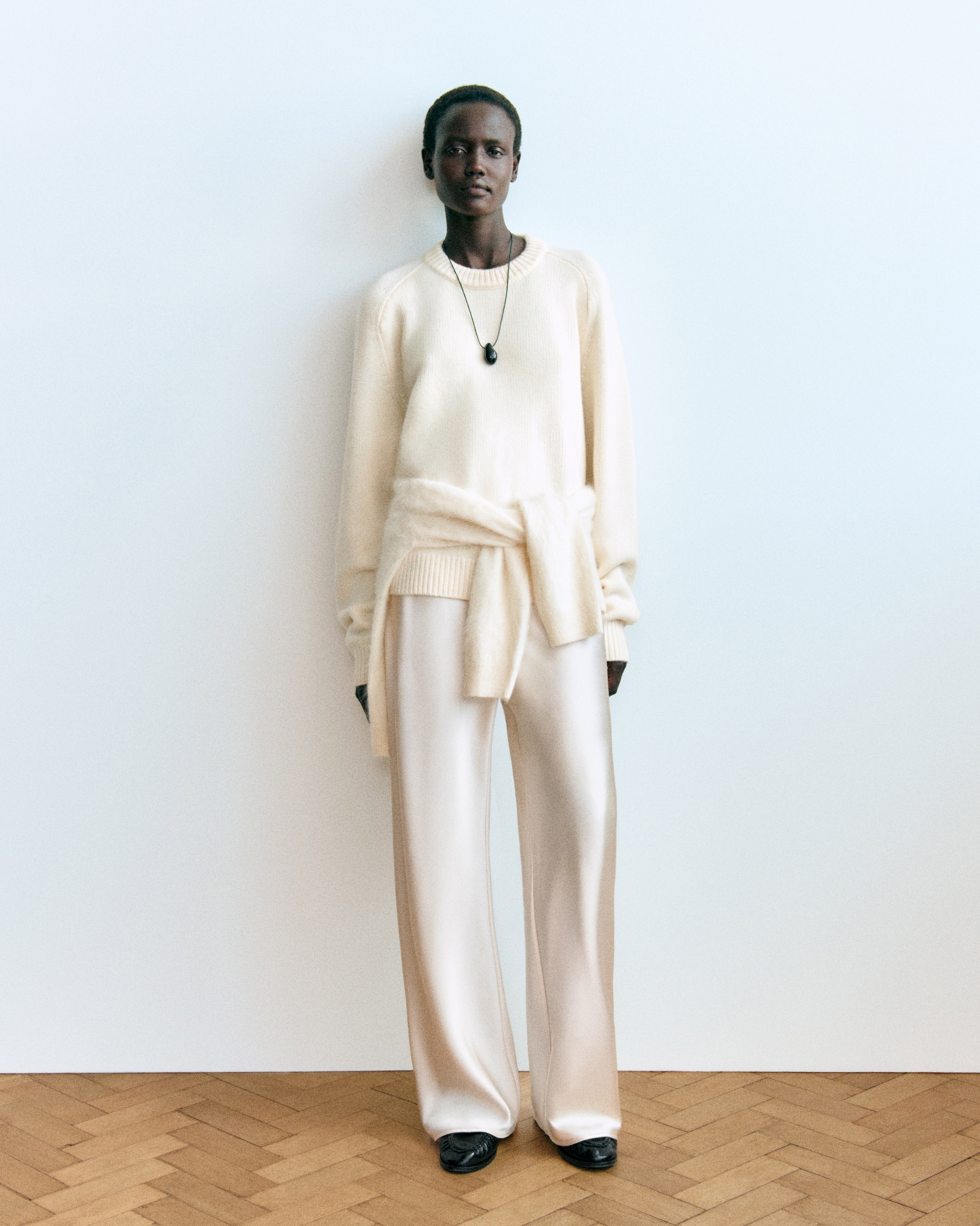 A person stands against a plain background wearing a cream sweater, cream pants, and black shoes with a long necklace.