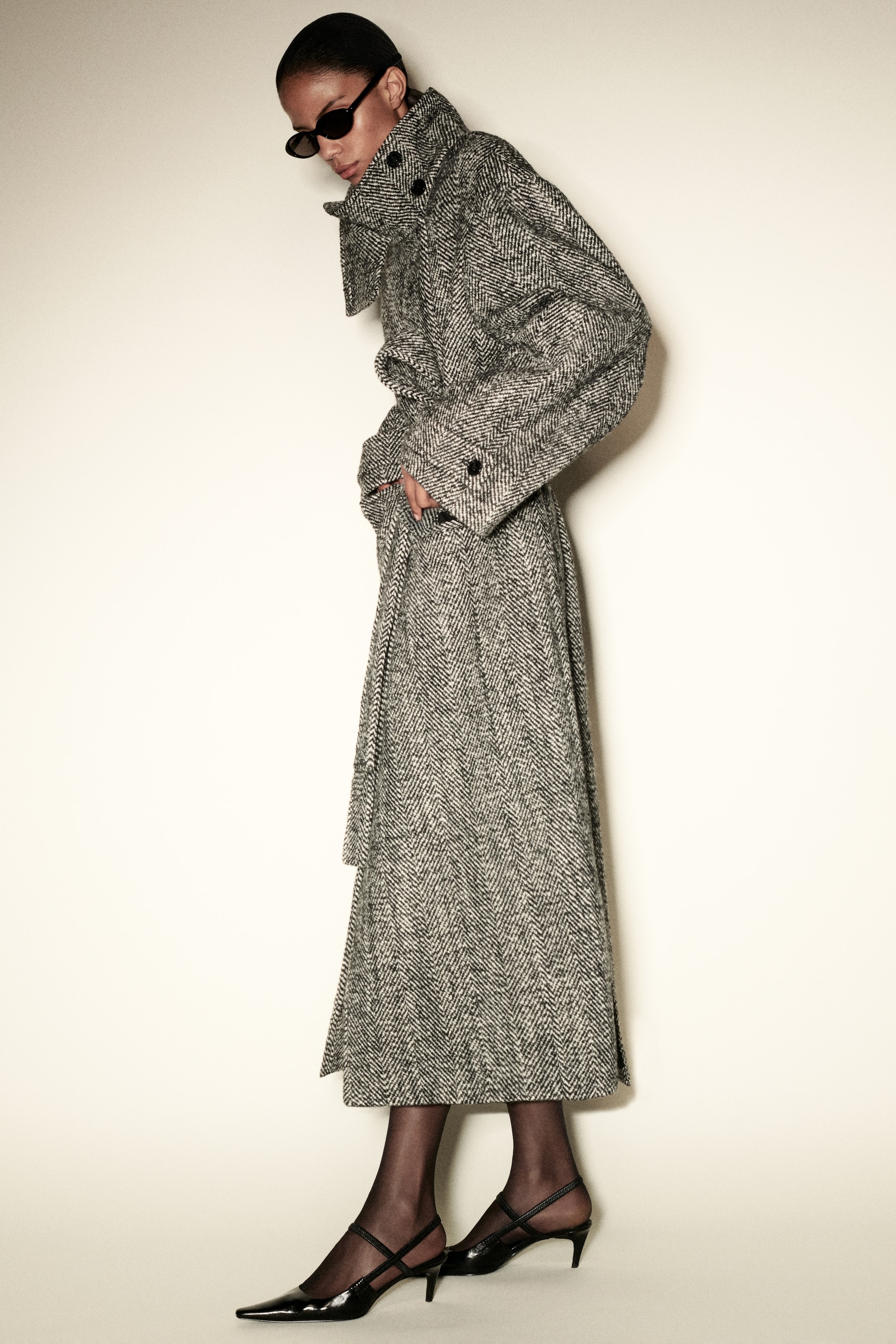 A person wearing a long gray herringbone trench coat, black heels, and sunglasses, posing against a plain background.