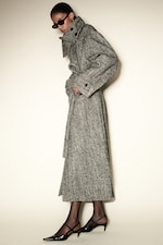 Placeholder for: A person wearing a long gray herringbone trench coat, black heels, and sunglasses, posing against a plain background.