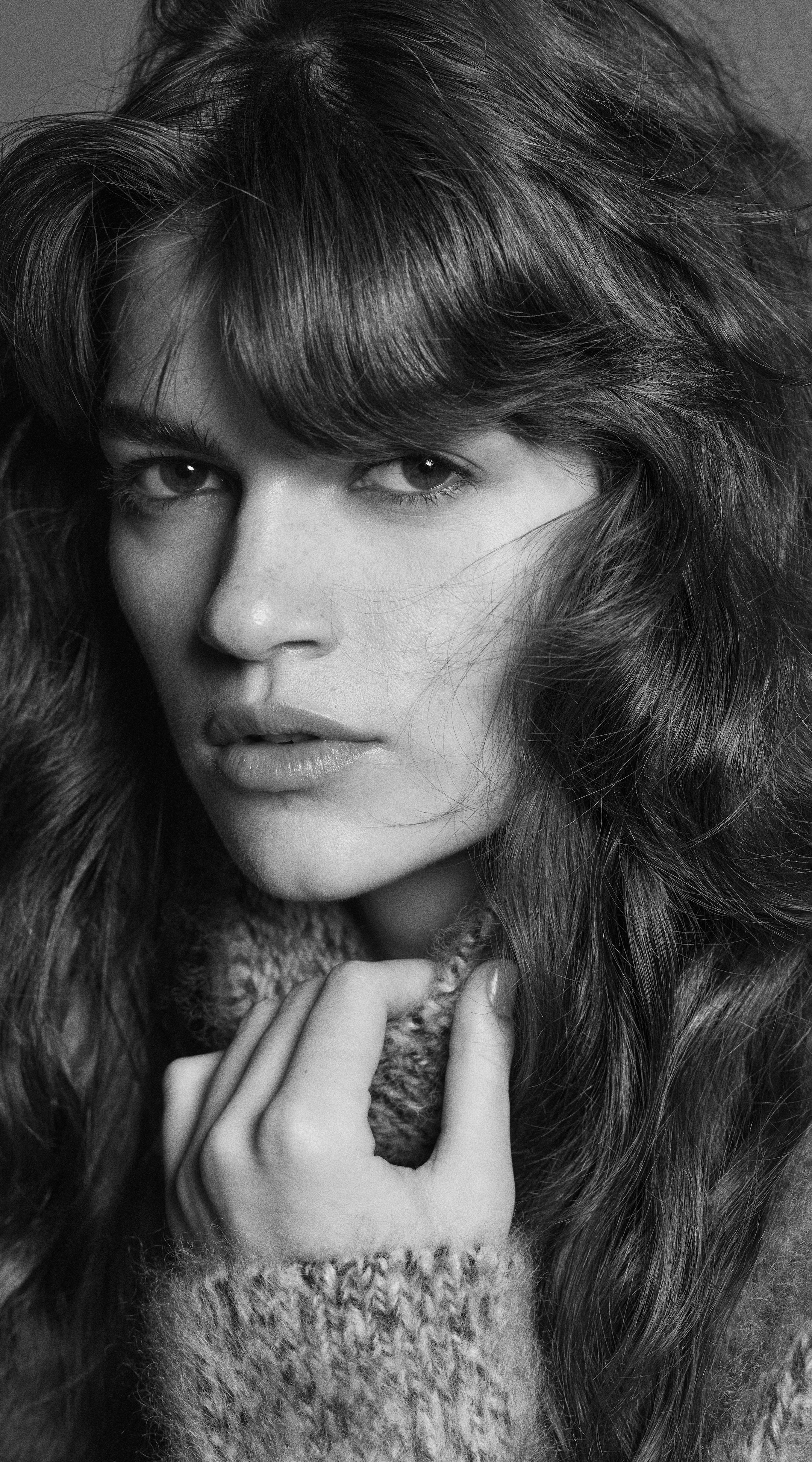 Black and white close-up portrait of a person with wavy hair, wearing a cozy sweater, gazing directly at the camera.