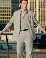 Placeholder for: Person in a light gray suit with an open collar shirt stands outdoors by a railing, with buildings in the background.