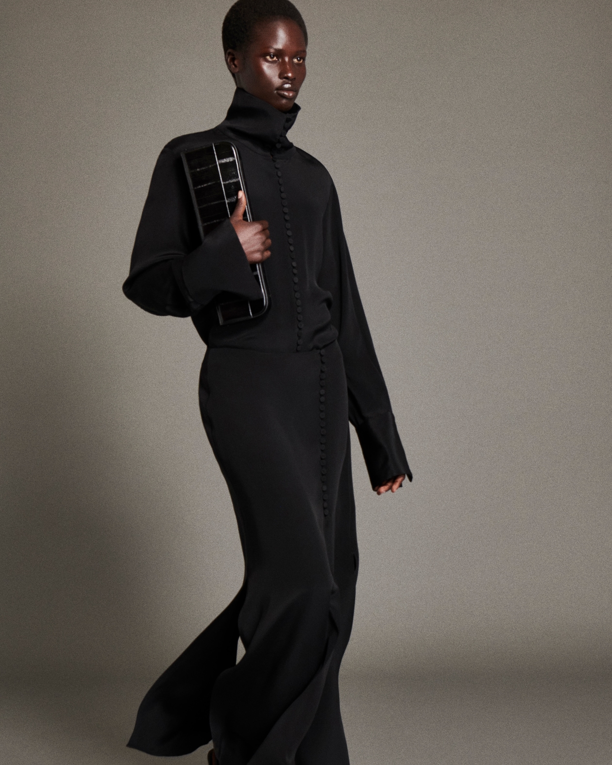 Person in a black outfit holding a black clutch, walking against a plain background.