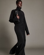 Placeholder for: Person in a black outfit holding a black clutch, walking against a plain background.