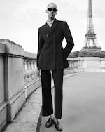 Placeholder for: A person in a tailored black suit and sunglasses stands on a bridge near the Eiffel Tower, in a stylish black and white photo.