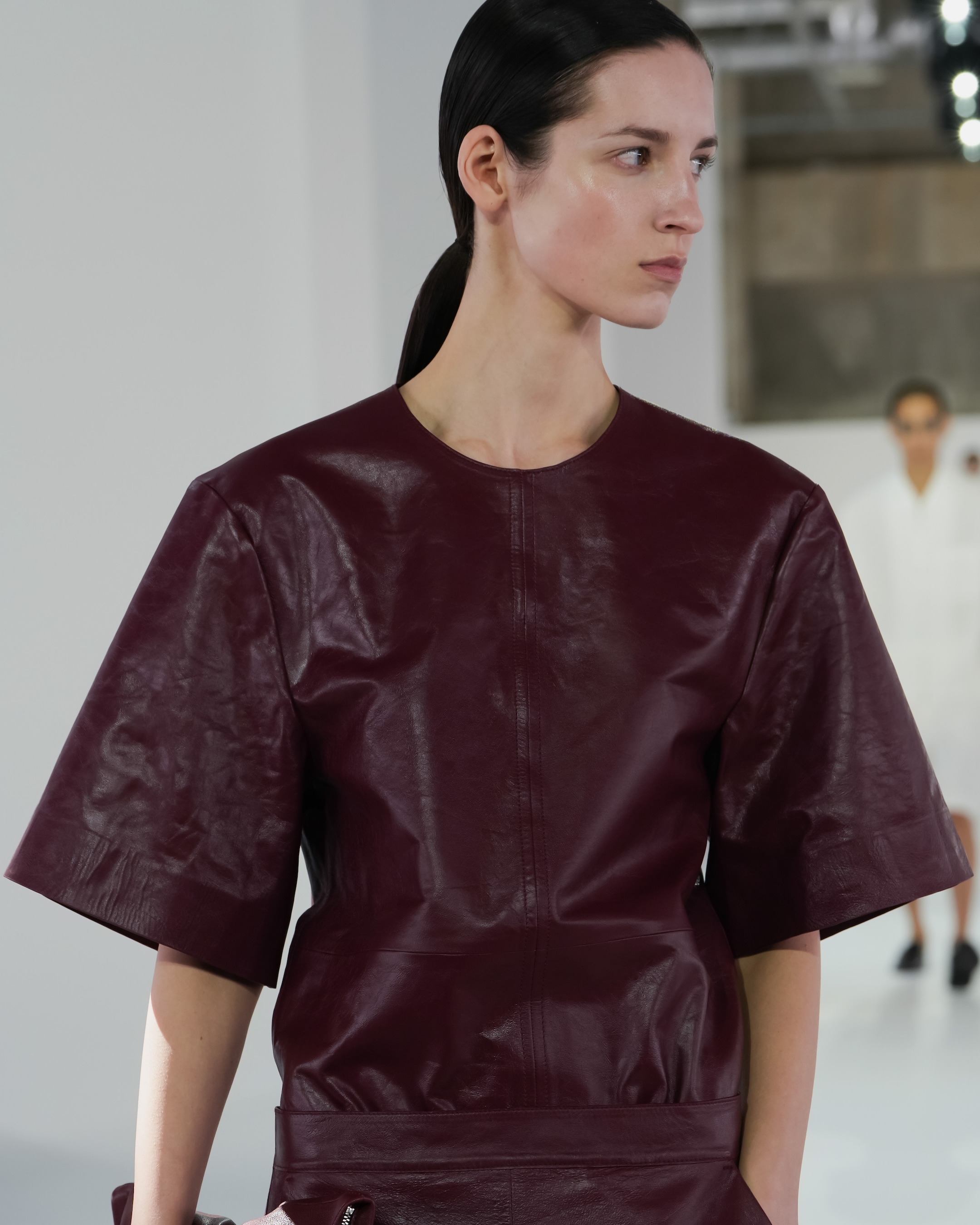 Model in dark red leather top and skirt with elbow-length sleeves walks on fashion runway, white background and second model blurred in distance.