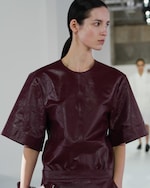 Placeholder for: Model in dark red leather top and skirt with elbow-length sleeves walks on fashion runway, white background and second model blurred in distance.