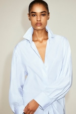 Placeholder for: A person with short hair wears an oversized, light blue, pinstripe shirt with an open collar, gazing confidently at the camera against a neutral background.