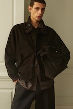 Placeholder for: A man wearing a dark suede jacket and shirt, holding fabric, stands against a neutral-colored wall with a confident expression.