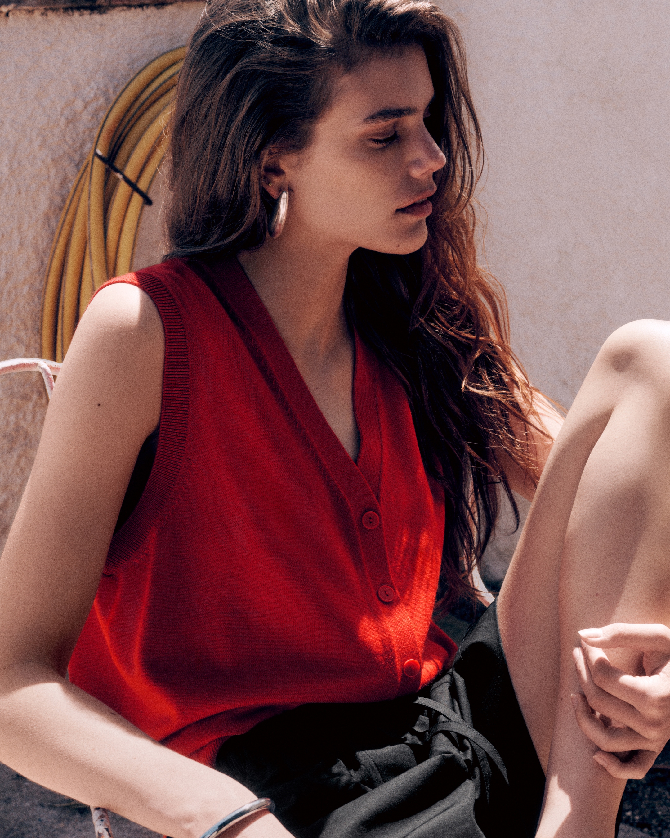 Woman in red vest cardigan and large gold hoop earrings, with black shorts on facing right