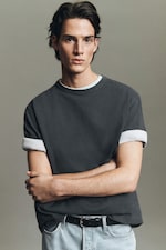 Placeholder for: Person with dark hair, wearing a dark rolled-up T-shirt and light jeans, standing with folded arms in front of a neutral background.