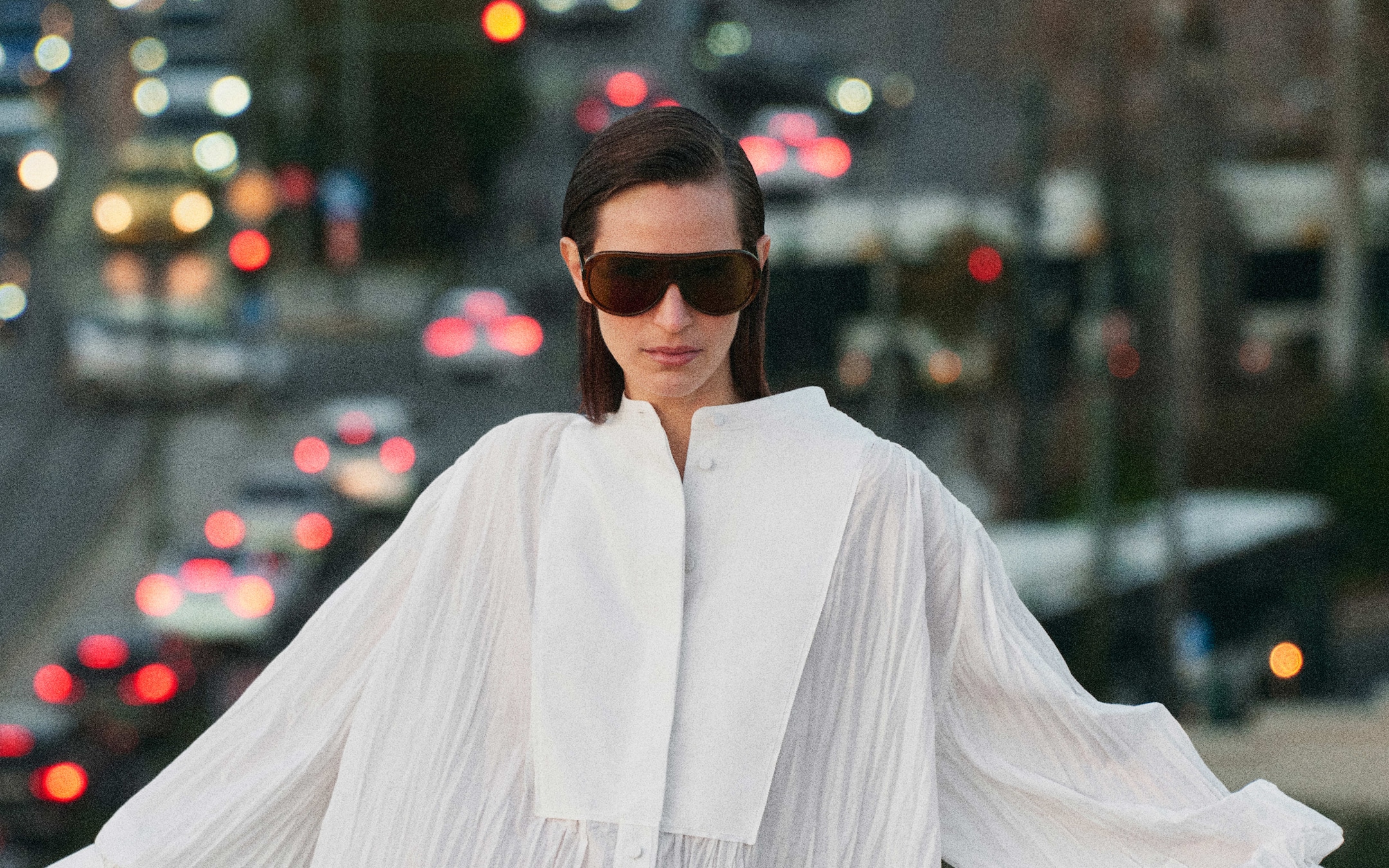 Person in white outfit and large sunglasses, standing against a blurred city background with bokeh lights from traffic.