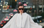 Placeholder for: Person in white outfit and large sunglasses, standing against a blurred city background with bokeh lights from traffic.
