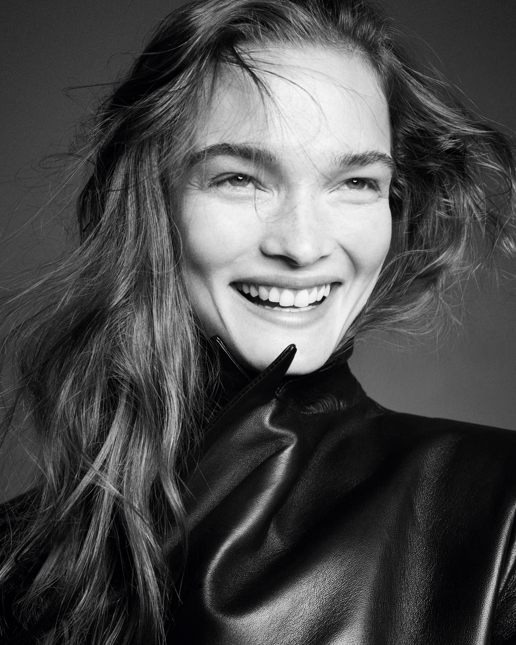 Woman smiling in a black and white portrait, wearing a leather jacket, with wavy hair and natural lighting.