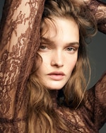 Placeholder for: Woman with long wavy hair poses with hands above her head, wearing a brown lace top, against a dark background.