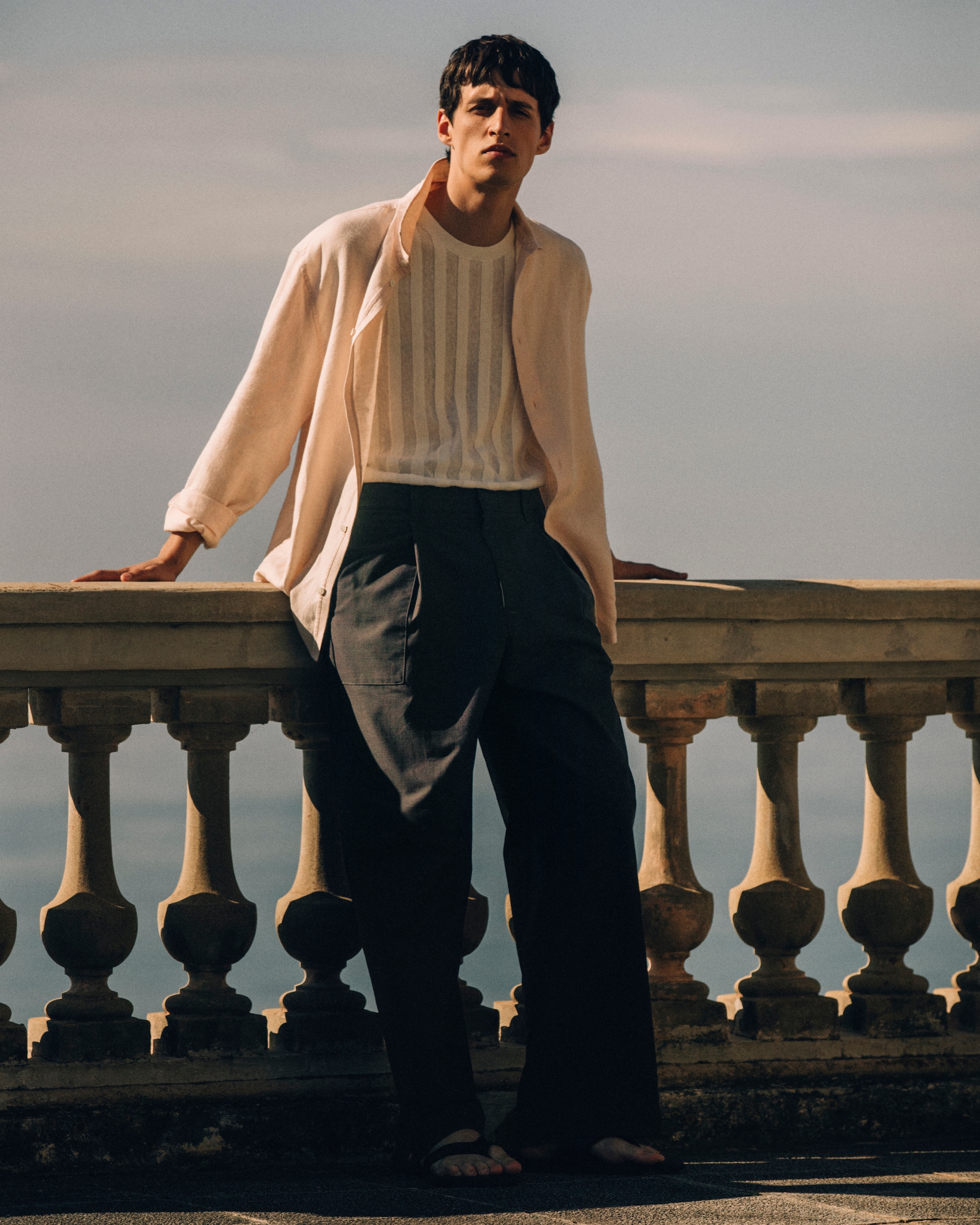 Man in striped knitted top and overshirt leaning on a stone balcony