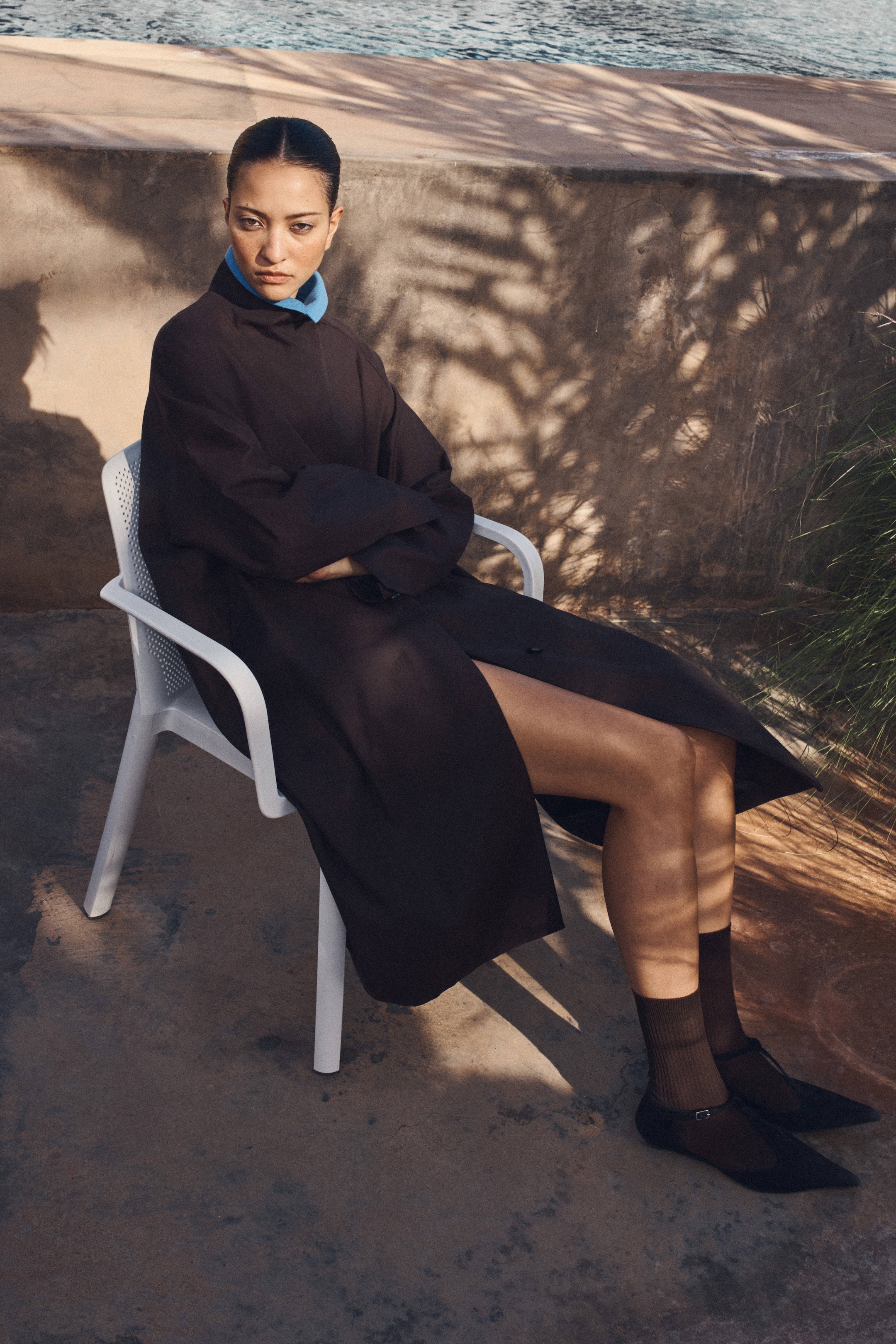 A person sits outdoors on a white chair, wearing a long dark coat, blue collared shirt, and black shoes, with a serious expression