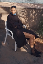 Placeholder for: A person sits outdoors on a white chair, wearing a long dark coat, blue collared shirt, and black shoes, with a serious expression