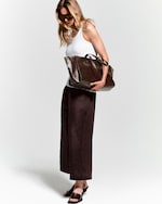 Placeholder for: Woman in sunglasses wearing a white tank top and wide brown pants holds a large brown bag against a plain background.