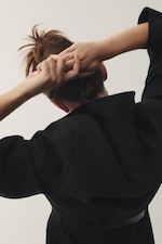 Placeholder for: Person in a black coat seen from behind, tying up their hair against a plain background.